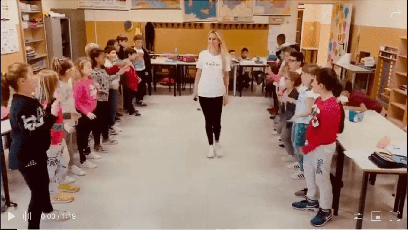 Allegra Lite teaching first year middle school children who are standing in two lines facing each other happy and ready to sing. Allegra is in the middle smiling and holding a microphone